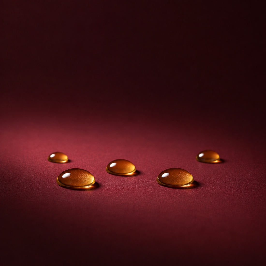 Des graines de jojoba à côté de trois gouttes ambrées d’huile de jojoba sur un fond bordeaux, un ingrédient du gloss à lèvres Bella Gloss