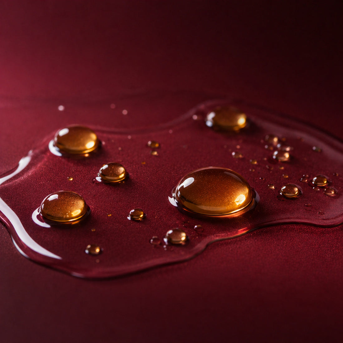 Trois capsules ambrées de vitamine E, dont une ouverte avec une goutte d’huile, sur un fond bordeaux, un ingrédient du gloss à lèvres Bella Gloss