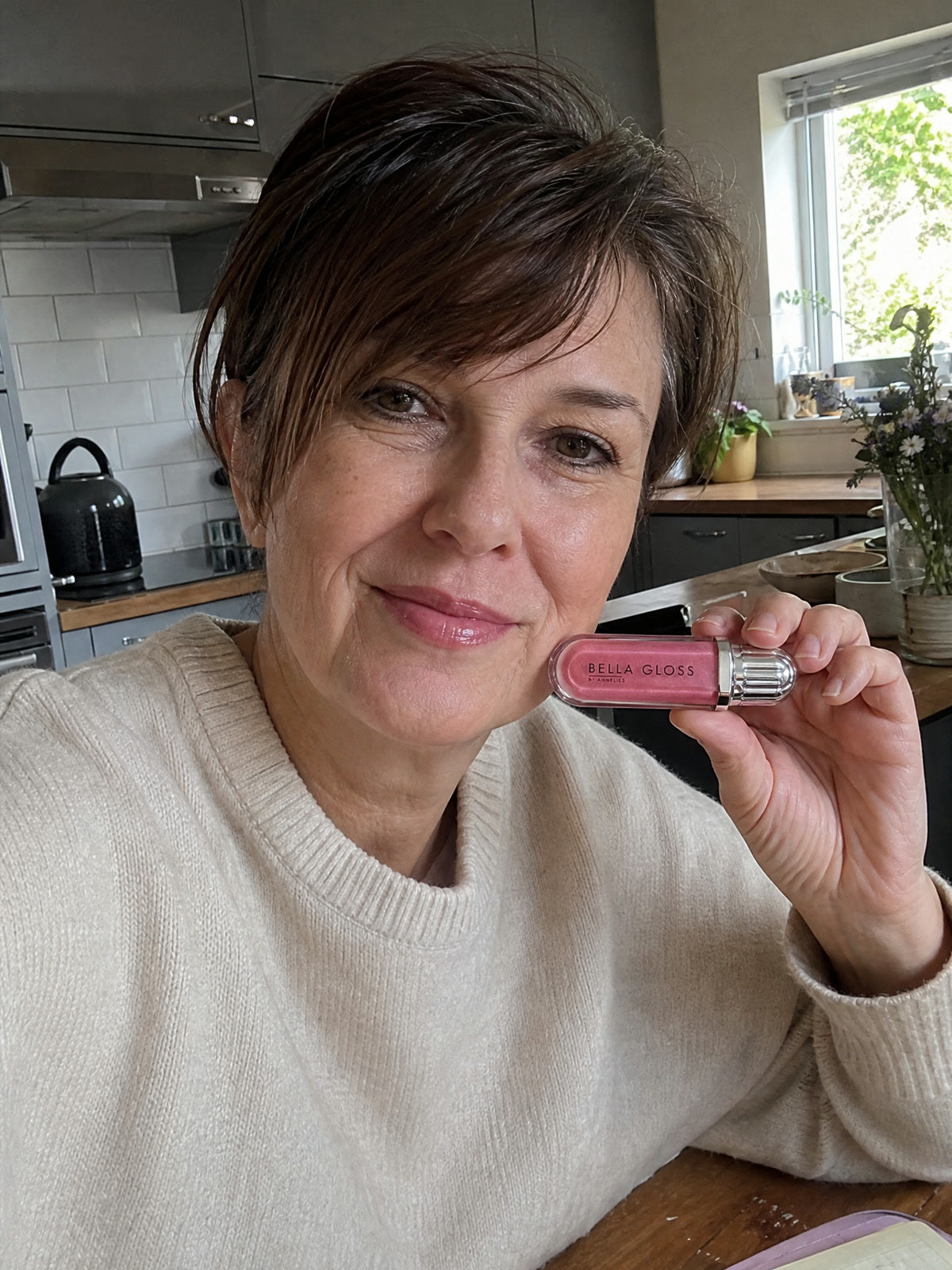 Une femme assise à la table de la cuisine lève le gloss à lèvres Bella Gloss