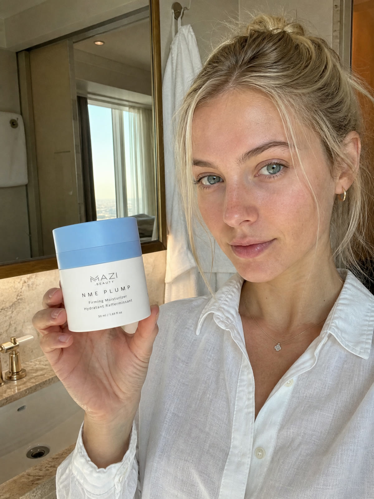 Une femme en chemise blanche tient le pot fermé de crème NME Plump dans sa salle de bain