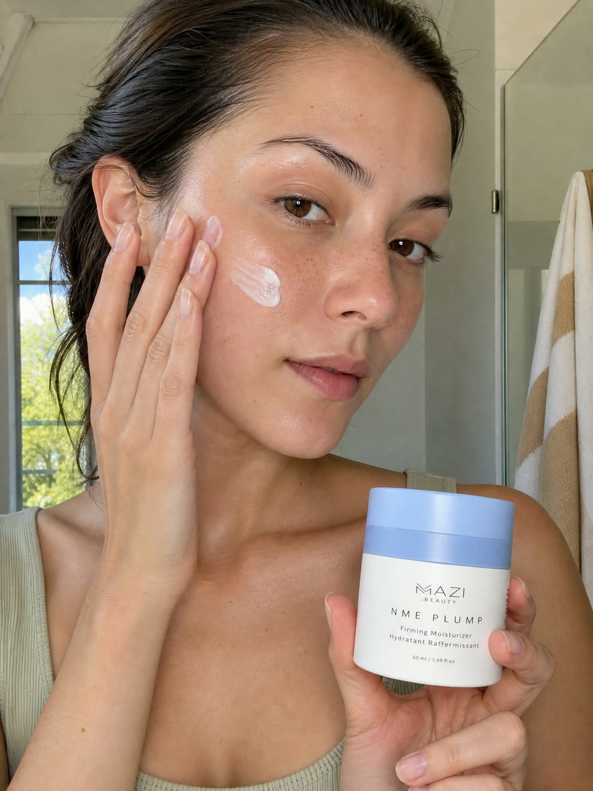 Une femme applique la crème blanche NME Plump sur sa joue avec les doigts, le pot dans l'autre main