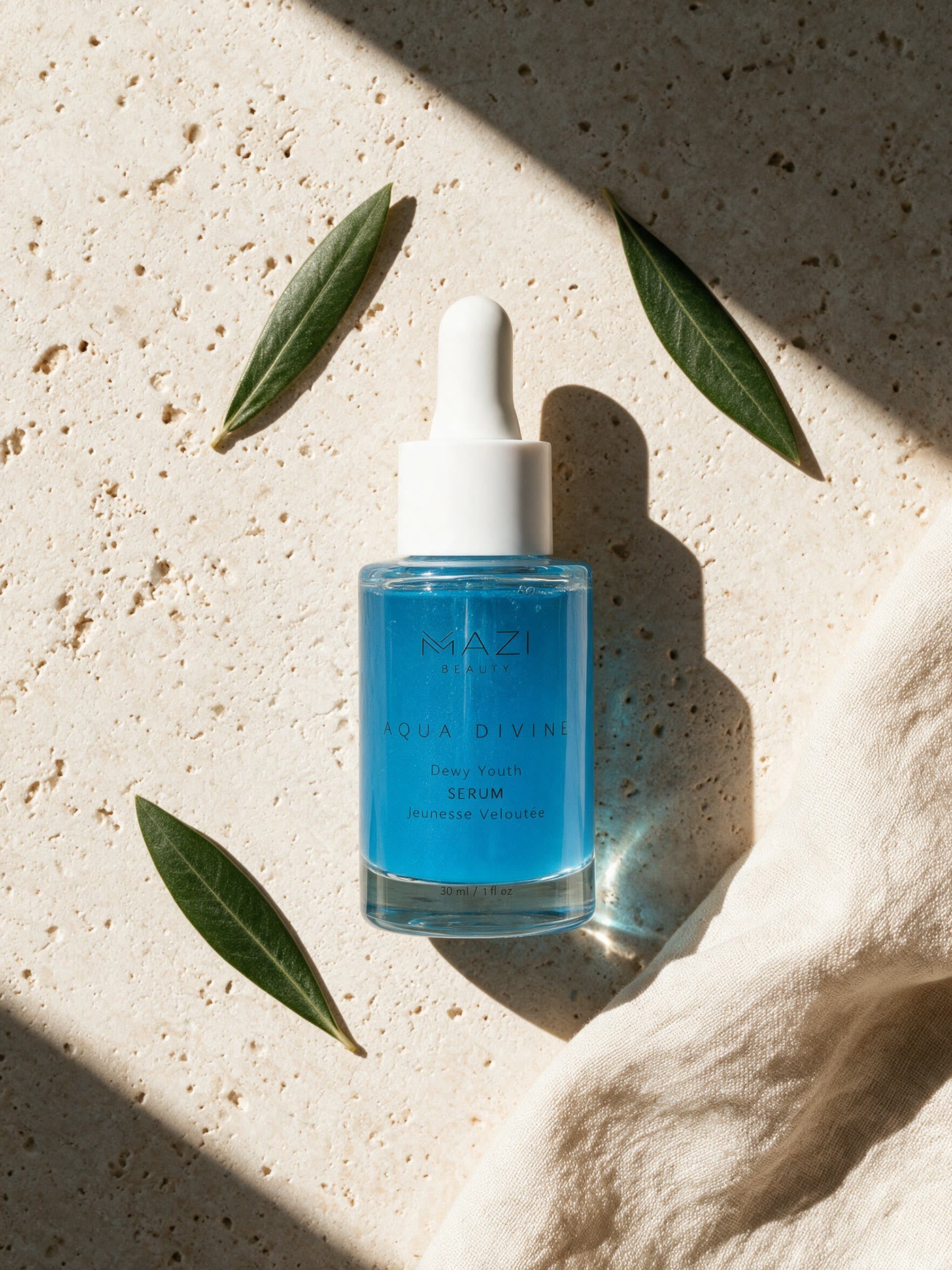 The closed Aqua Divine serum bottle photographed from above, lying on travertine between olive leaves and linen