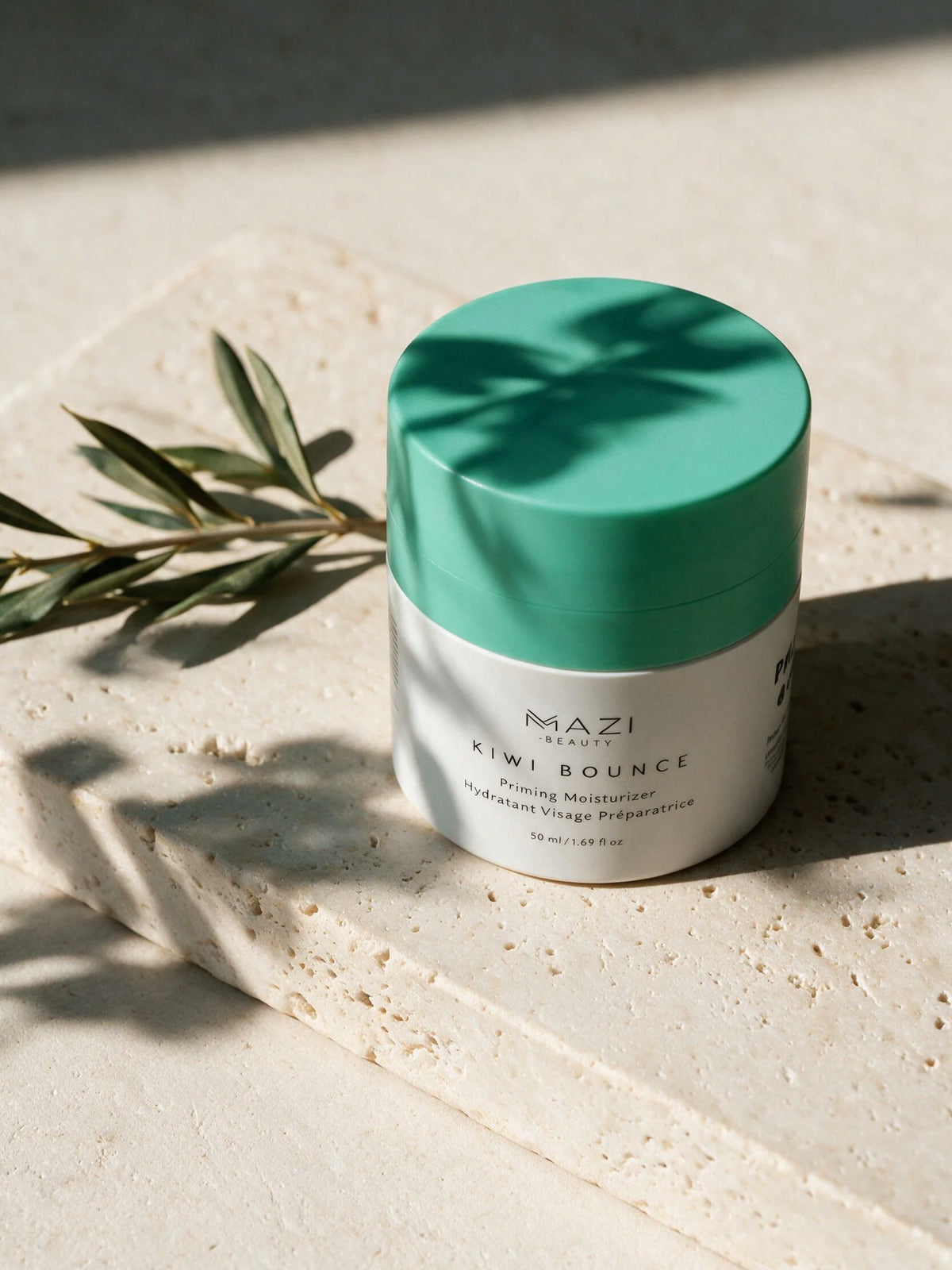 The closed Kiwi Bounce jar on travertine beside an olive branch in hard daylight