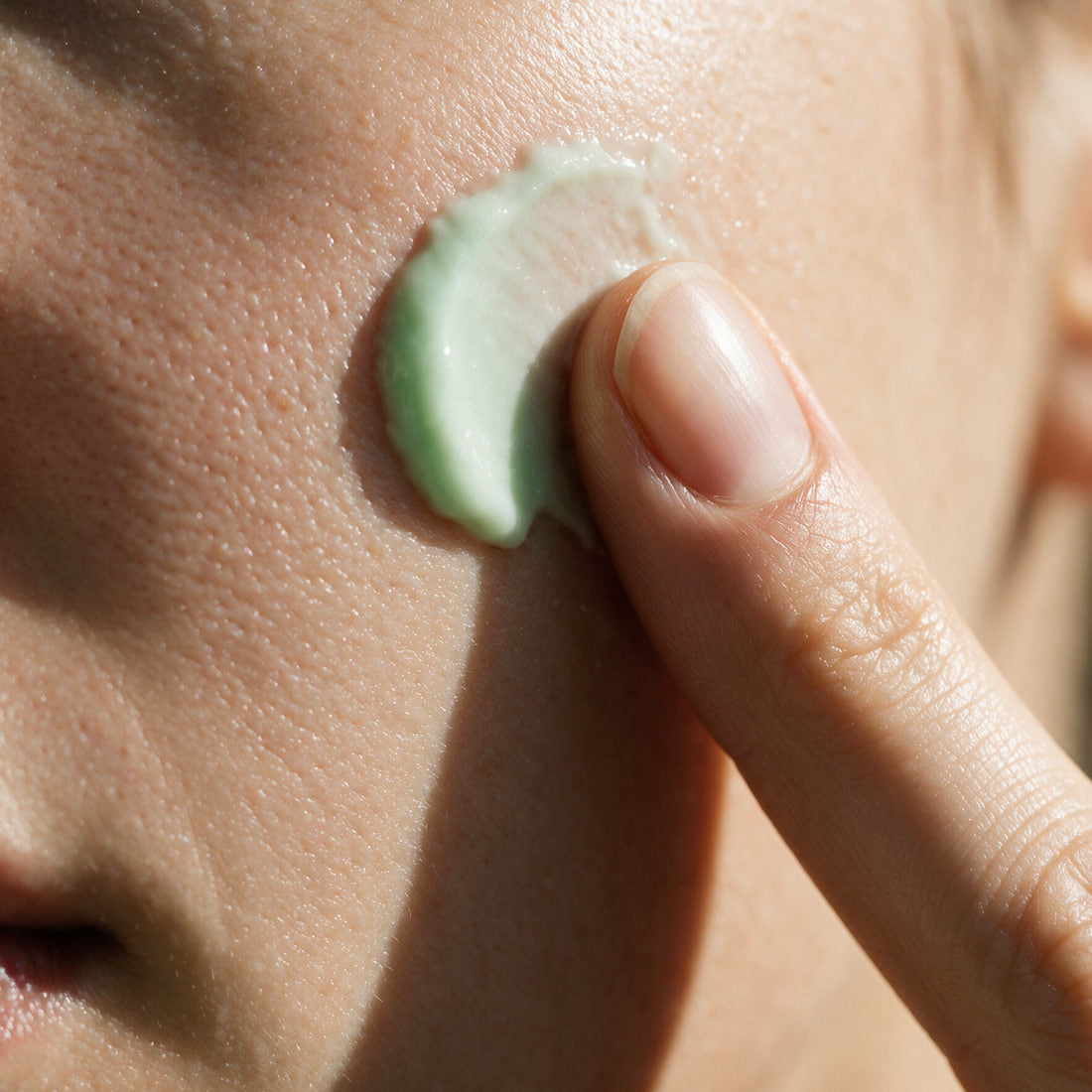 A fingertip carrying pale green Kiwi Bounce day cream touches a woman's cheek