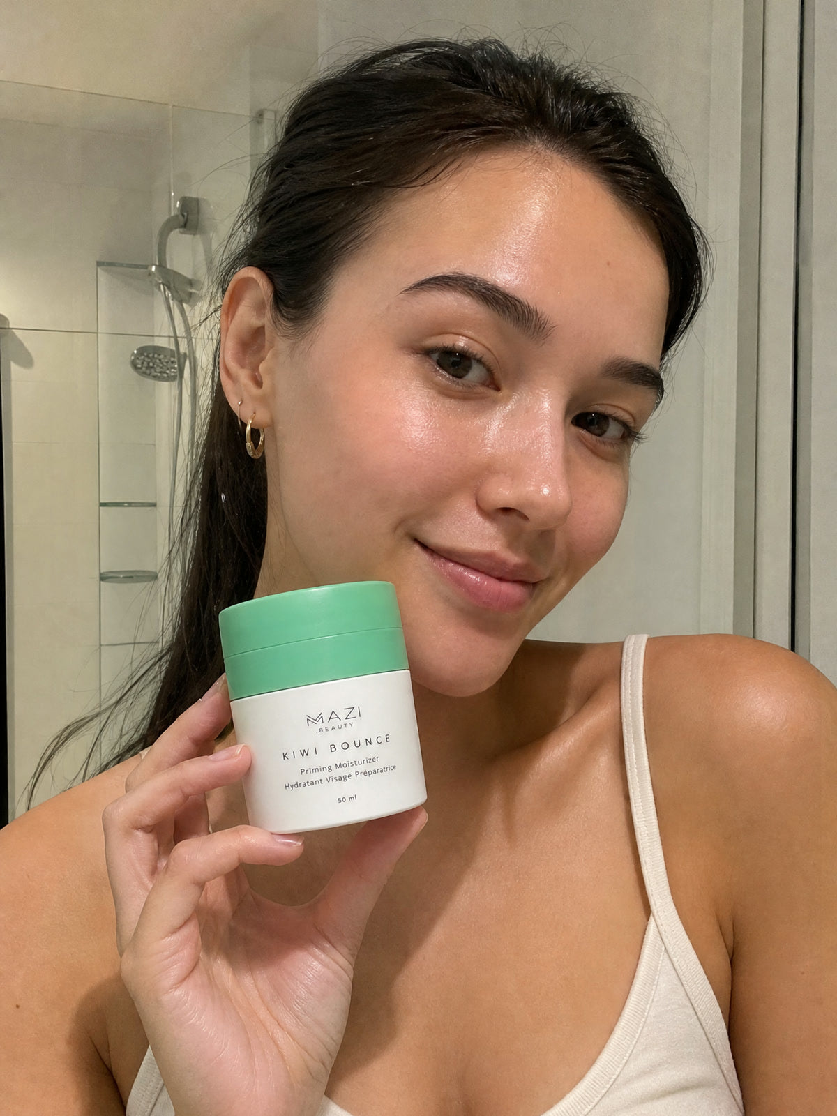 A woman in a white vest holds the Kiwi Bounce day cream jar in her bathroom, the shower behind her