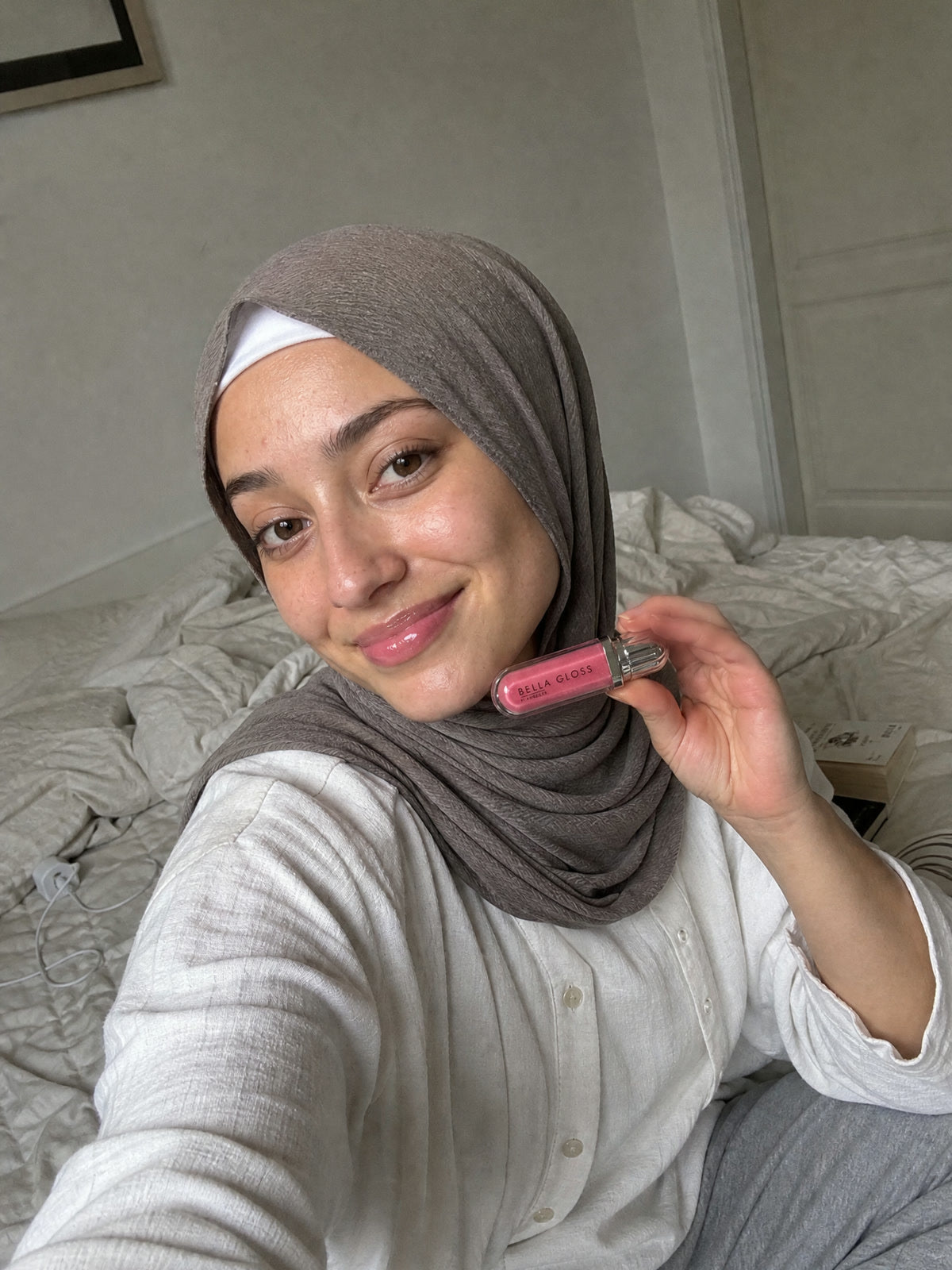 Une femme portant un foulard est assise sur un lit avec le gloss à lèvres Bella Gloss