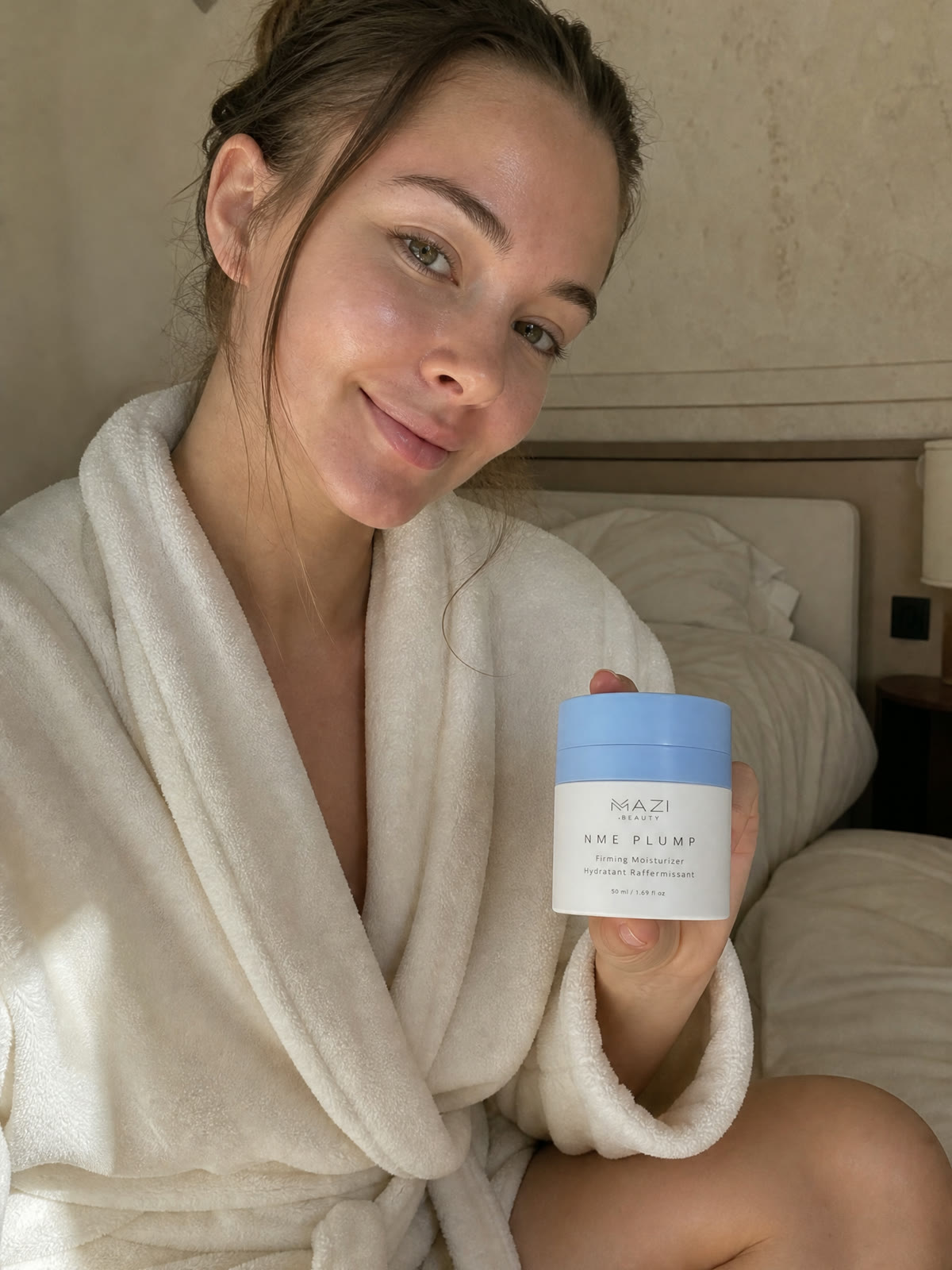 Une femme en peignoir blanc tient le pot fermé de crème NME Plump au bord de son lit