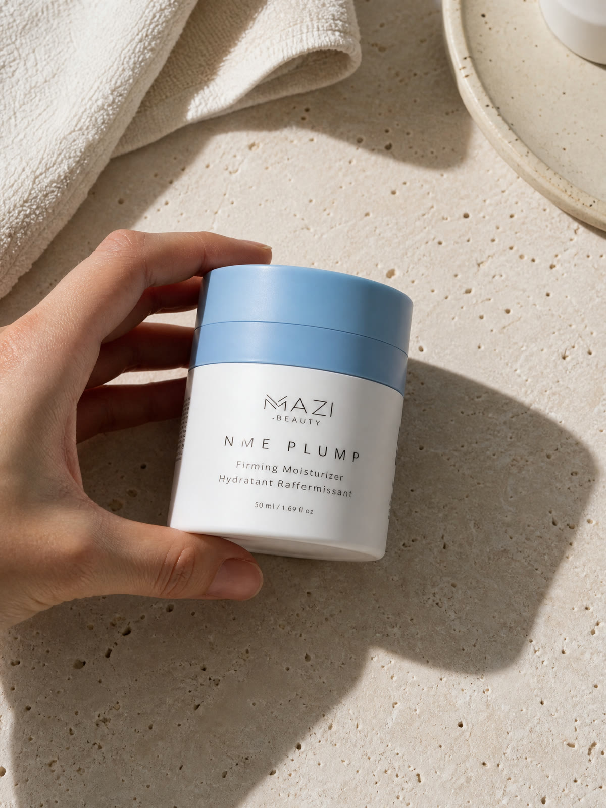 Une main tient le pot fermé de crème NME Plump au-dessus d'un plan en travertin, à côté d'une serviette