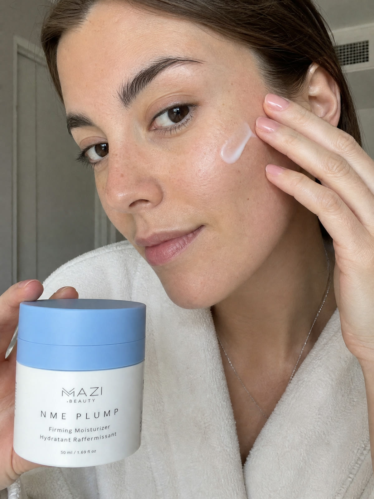 Une femme avec une noisette de crème NME Plump sur la joue tient le pot fermé devant elle