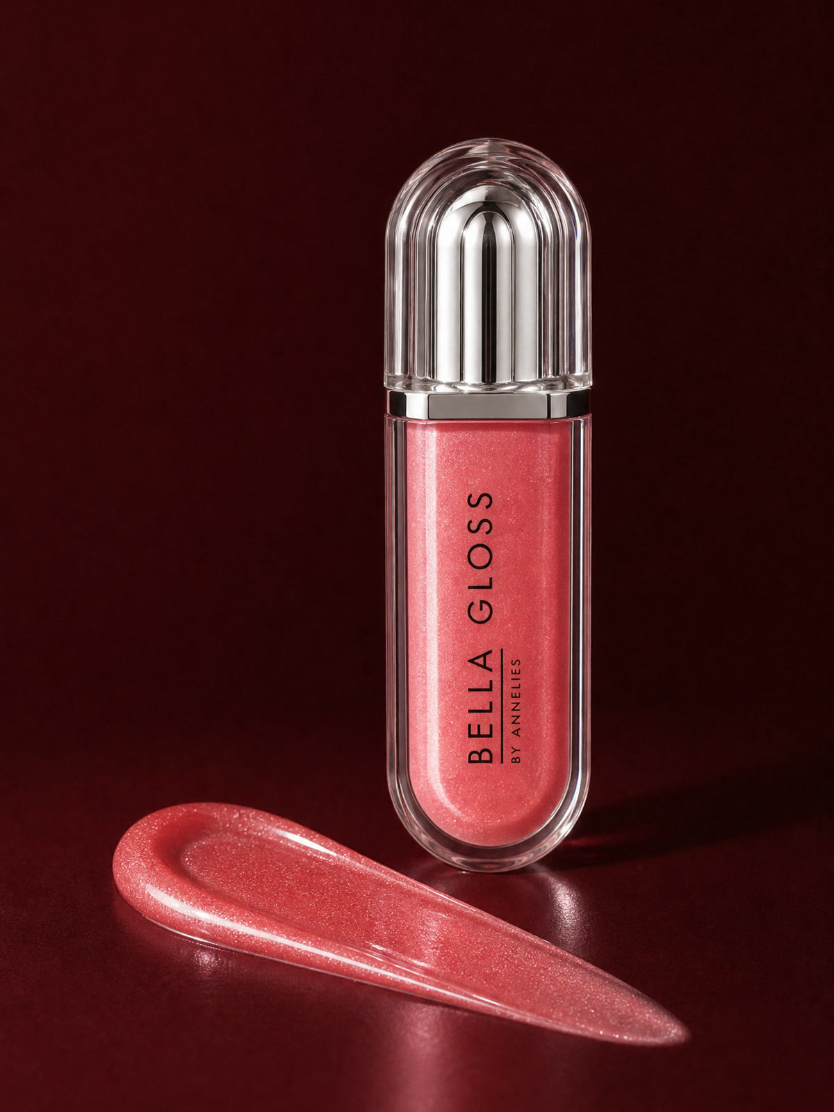 The closed coral-red Bella Gloss lip gloss tube stands upright beside a glossy swipe of product