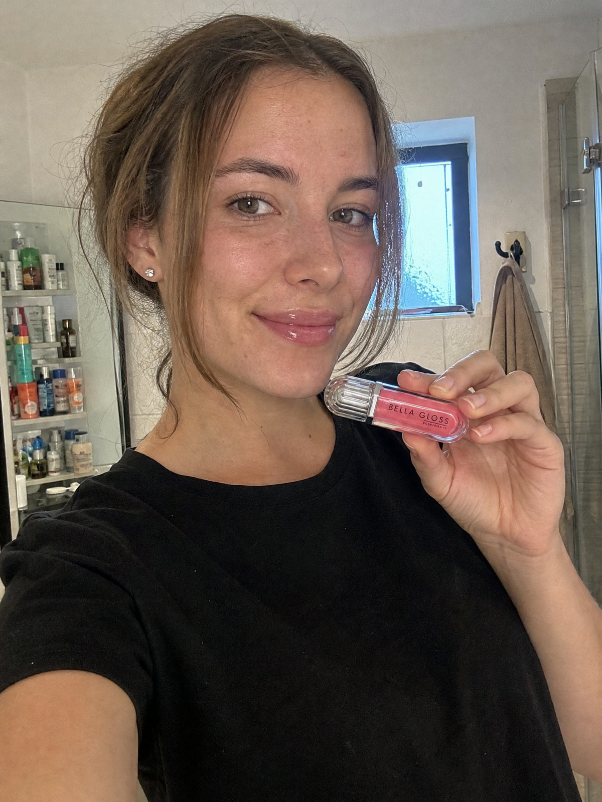 A woman shows Bella Gloss lip gloss in front of the bathroom mirror